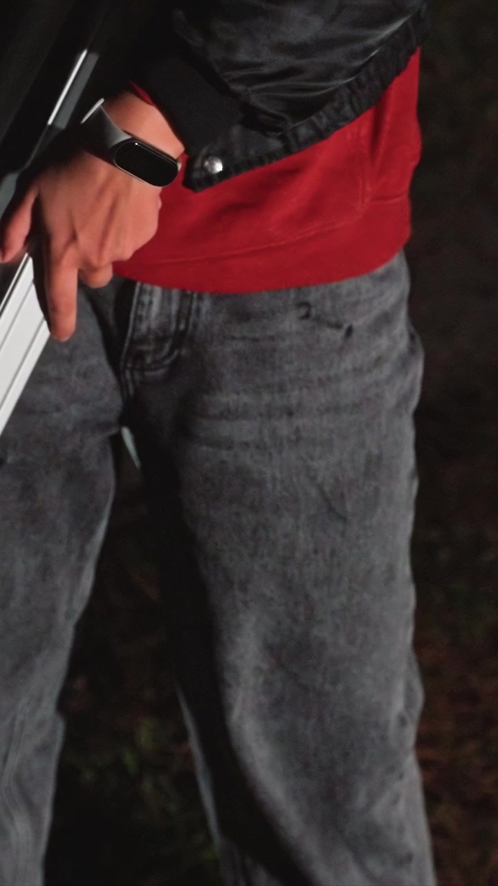 Man in red shirt gray pants and black jacket holds vehicle. Electronic wristpiece is evident on person appendage. Low light setting outdoors highlights subject midsection