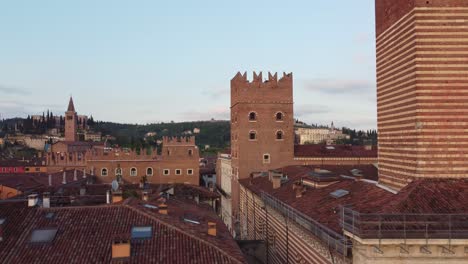 descending drone shows verona palazzo del podesta and crenellated torre dei lamberti with striped palazzo della ragione on the right, rooftops and hillside churches visible, real time, golden hour