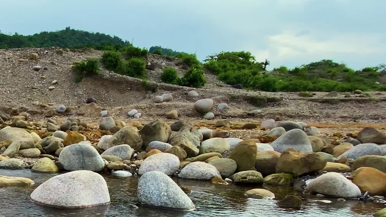 Bulutlu bir gökyüzünün altında, çakıllı bir setin ve yeşil bir tepenin yanından, pürüzsüz kayaların ve çakılların etrafından akan sığ bir derenin alçak açılı görünümü.