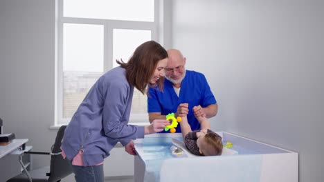 a confident man with a gray beard and glasses an experienced pediatrician in a blue doctors uniform plays with a little girl as a baby checking her developmental norms at her age in a modern clinic together with the girls mother