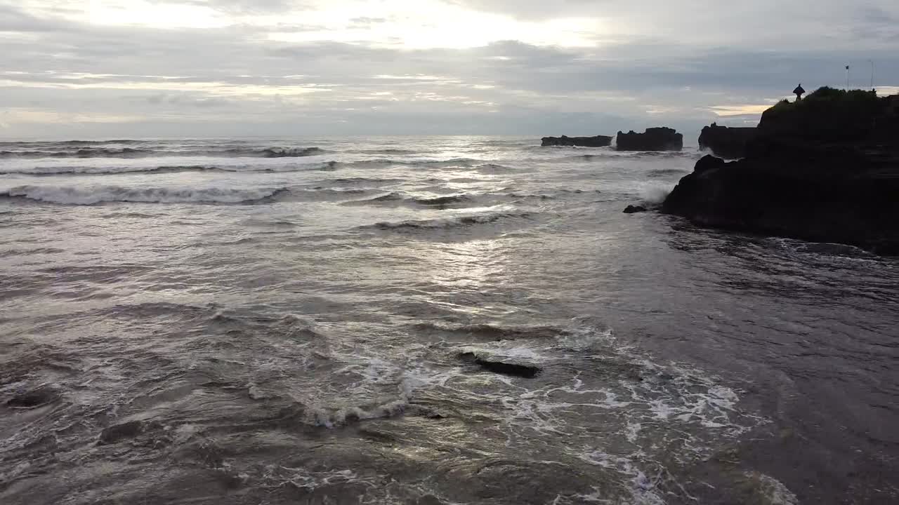 Ripresa aerea di onde oceaniche che si infrangono sulla costa rocciosa a Tanah Lot, Bali, Indonesia, con acqua texturizzata, riflessi tenui e cielo nuvoloso che creano un'atmosfera rilassante
