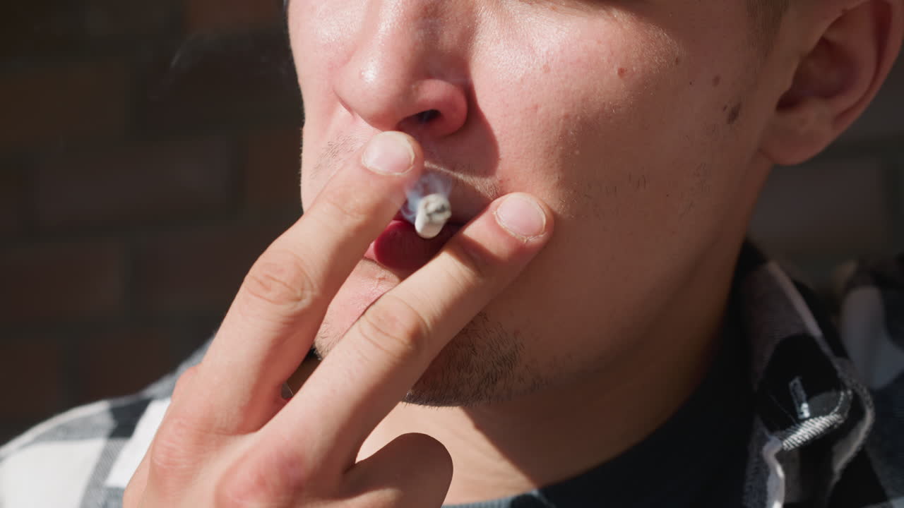 Close up young man in black white checkered flannel shirt holding lighter and cigarette between fingers, puffing smoke stream with brick wall and iron railing blurred in warm sunlight background