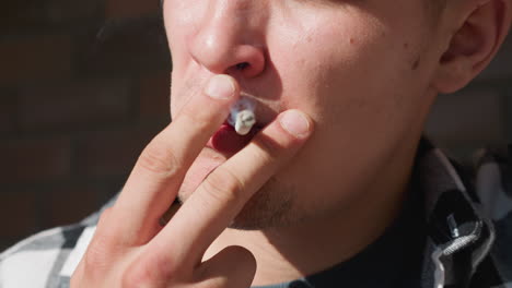 close up young man in black white checkered flannel shirt holding lighter and cigarette between fingers, puffing smoke stream with brick wall and iron railing blurred in warm sunlight background