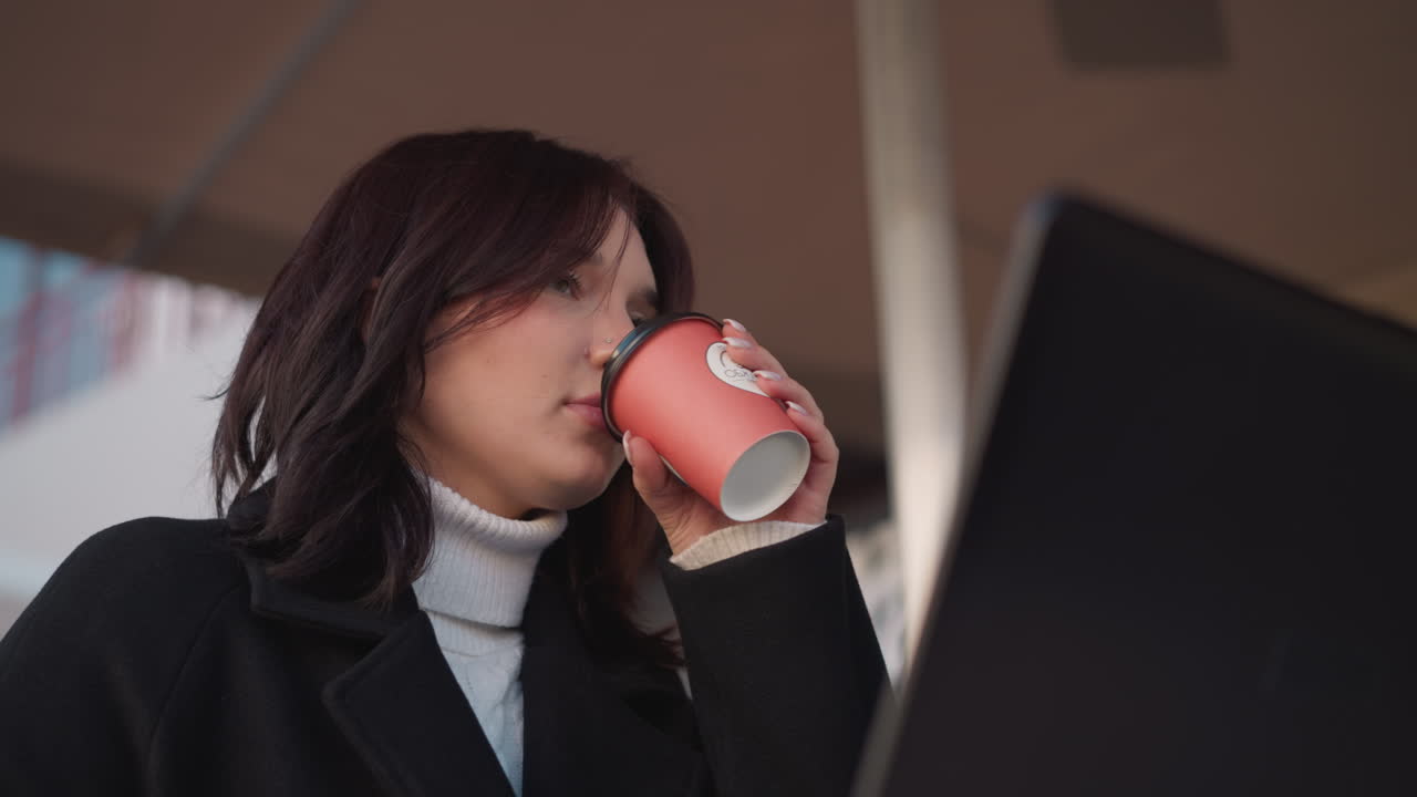Lady in black coat lifts tea cup under patio, pauses to sip, bursts into laughter, and gently places cup back on table, with a partial laptop visible in front