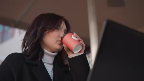 lady in black coat lifts tea cup under patio, pauses to sip, bursts into laughter, and gently places cup back on table, with a partial laptop visible in front