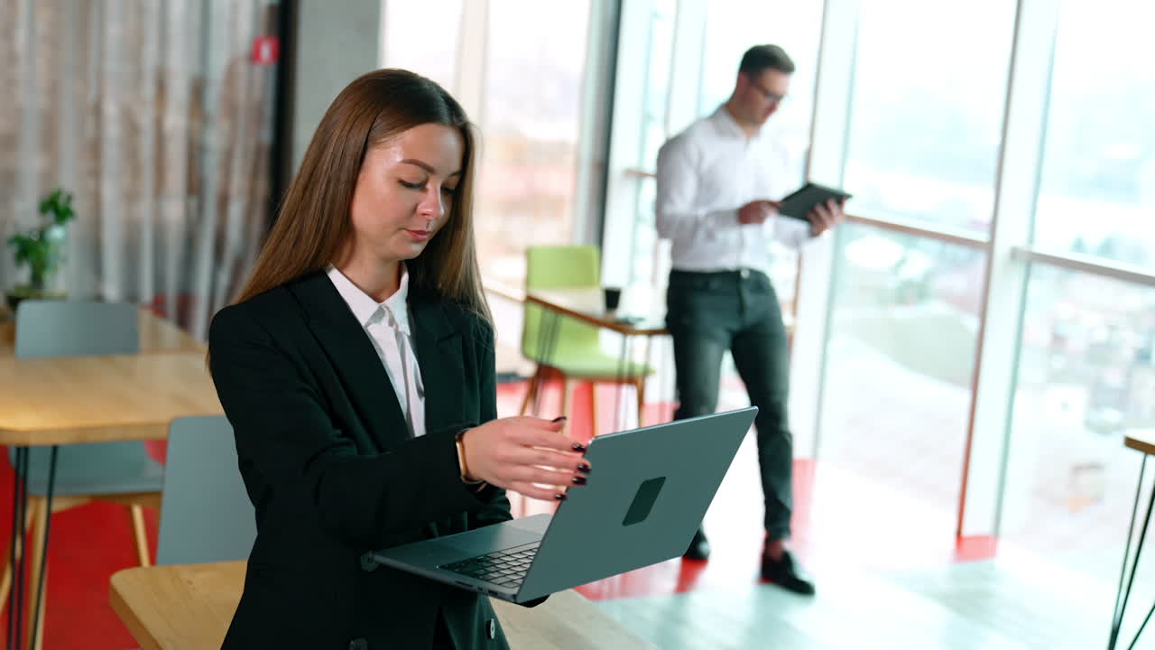 Successful modern business lady types on laptop and then closes it. Man at blurred backdrop works on I-pad.
