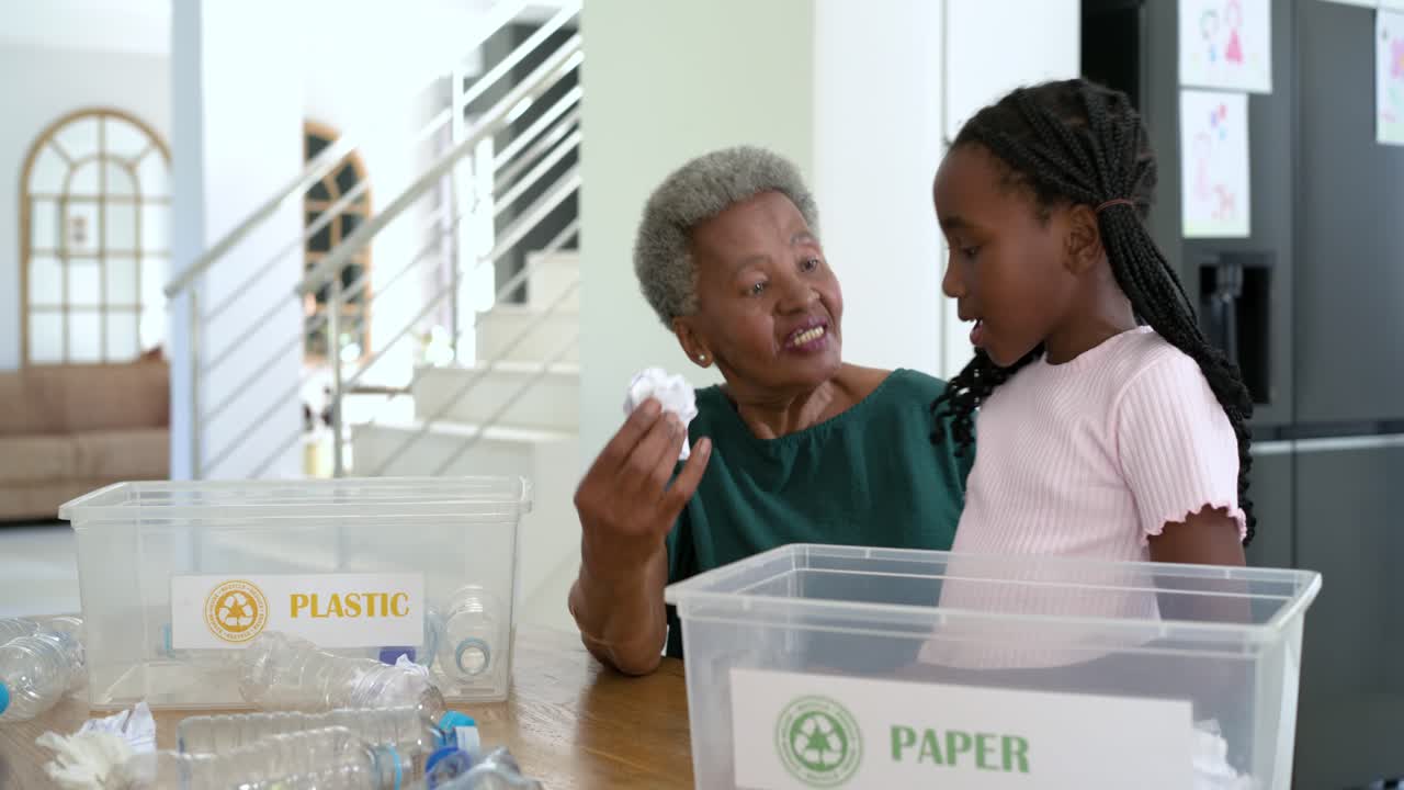 African American grandma guiding granddaughter sorting PAPER PLASTIC bins at table teaching sorting