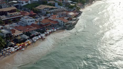 Rotating-aerial-drone-birds-eye-shot-of-the-beautiful-tropical-famous-Pipa-beach-during-high-tide-with-tourists-playing-in-the-water-and-enjoying-the-shade-under-colorful-umbrellas-on-a-summer-evening