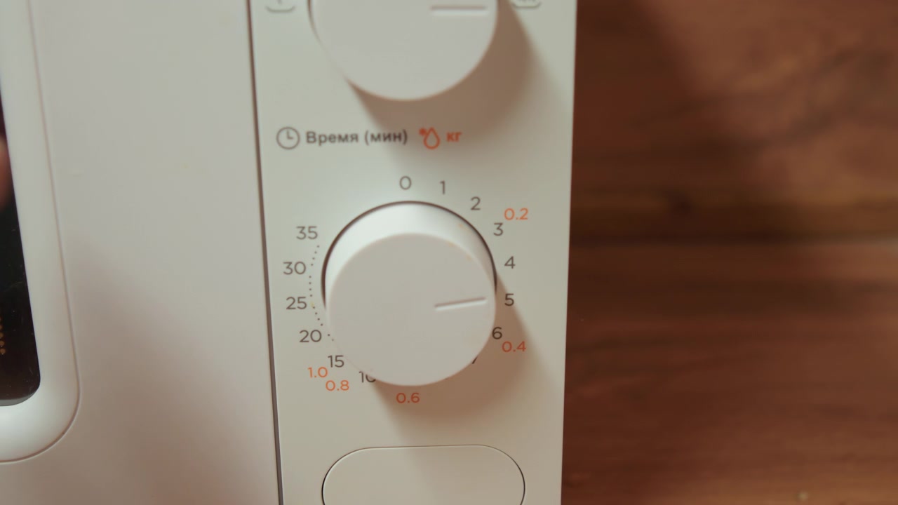 Una vista ravvicinata mostra una mano che gira la manopola inferiore del timer rotante sul pannello di controllo bianco di un forno a microonde in una cucina domestica, impostando il tempo di cottura accanto alle marcature stampate, mentre le dita creano una leggera sfocatura di movimento durante l'azione.