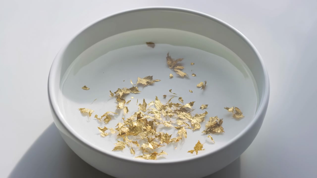 Vista ravvicinata dall'alto di una ciotola di ceramica bianca con scaglie di foglia d'oro galleggianti, leggere increspature sulla superficie, morbidi riflessi e uno stile minimale elegante su uno sfondo neutro e pulito.