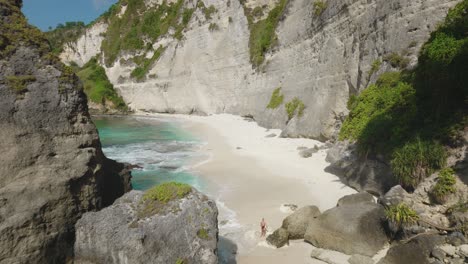 Playa-De-Diamantes-En-Nusa-Penida-Con-Acantilados-Escarpados-Y-Mujer-Corriendo-Entre-Rocas,-Antena