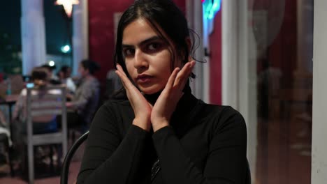 mujer joven en negro contemplando en un café durante la noche, con luces oscuras y clientes en el fondo