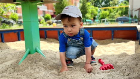 adorable caucasian baby sits in a sandbox. little cute kid in a cap first touches sand with hands and then takes toy rakes.