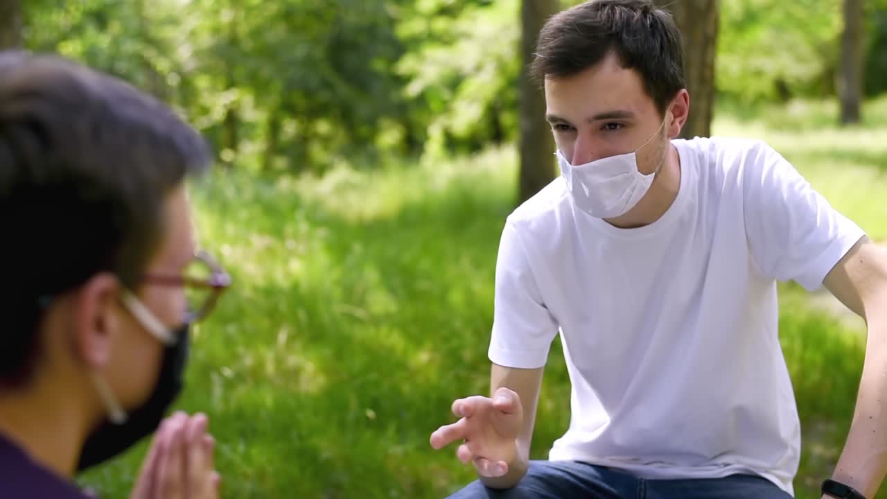 Two friends in protective medical masks talking in the park while resting on the bench by keeping their social distance from each other. Sunny day. Corona Virus idea