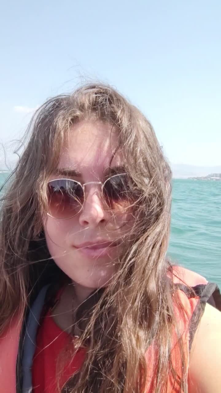 A woman with long hair and sunglasses is smiling for the camera, takes a selfie on a boat.