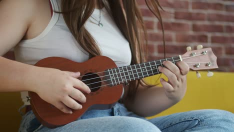 una mujer músico está sentada en el sofá y tocando el ukulele tocando cuerdas, vista frontal. una mujer hipster toca el ukulele sentada en un sofá