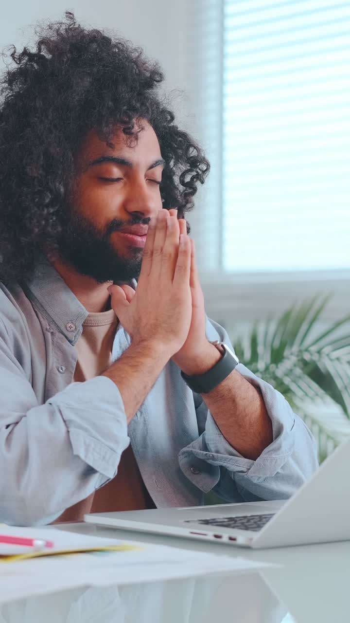 Positive arabian man freelancer makes praying gesture sits at desk with computer