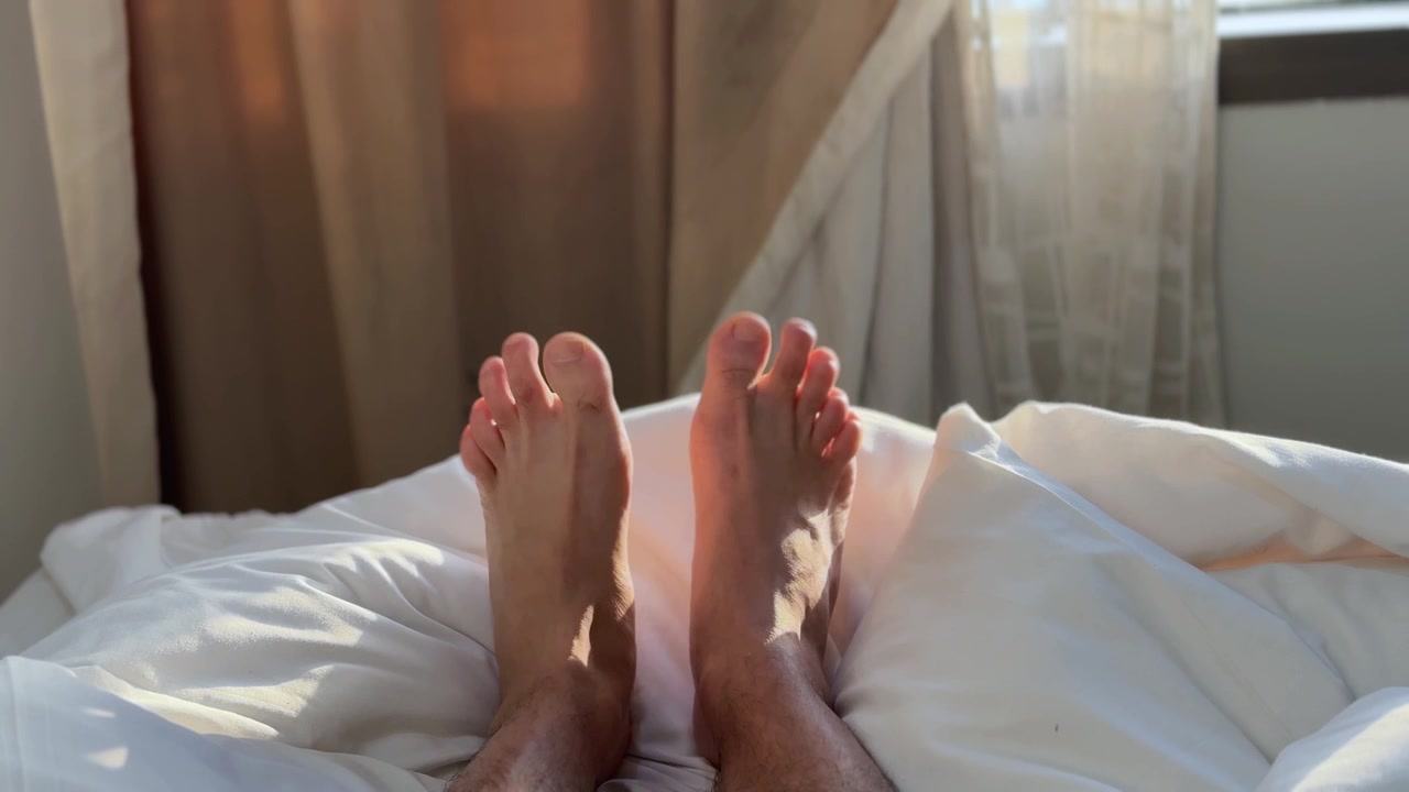 First-person view of bare male feet resting comfortably on soft white bed sheets. Bright morning sunlight shines through the bedroom curtains, illuminating the relaxing indoor vacation atmosphere.