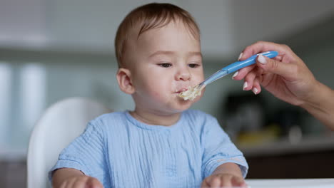 feeding a little kid from a spoon. mom's hand shoves a full spoon to baby's mouth. close up. blurred backdrop.