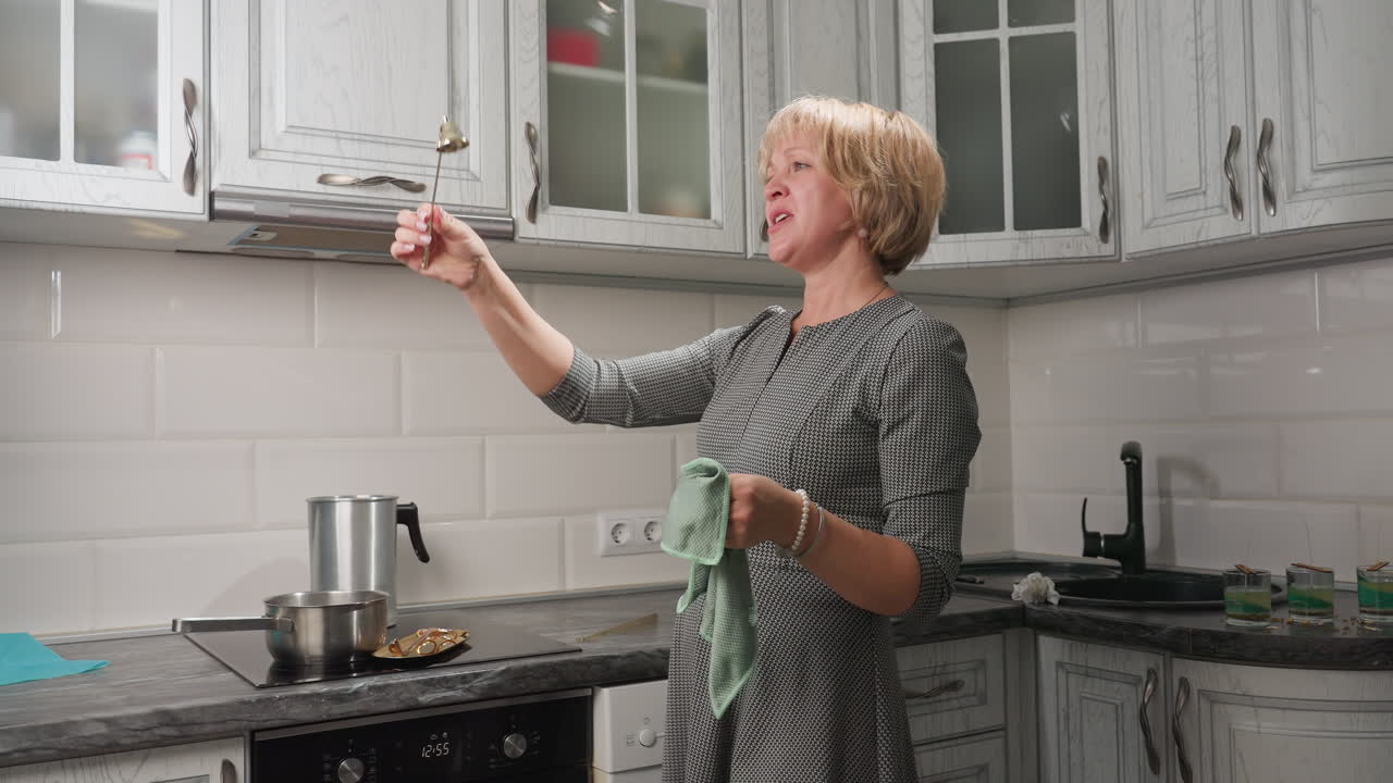 Female cake decorator in patterned dress joyfully sings and sways while examining golden bell decoration in bright kitchen with cabinets, oven, and candle molds lined on counter beside black sink