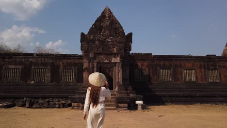 young beautiful woman in long white dress and vietnamese hat going to palace in wat phou ruined khmer hindu temple complex. champassak, laos, asia. sunny. ancient culture religious architecture. slow