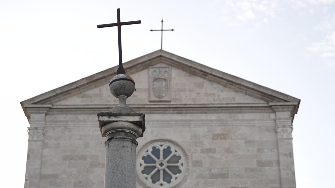 Coluna de pedra em forma de cruz em primeiro plano diante da fachada de uma antiga igreja romana com uma rosácea circular em um dia claro