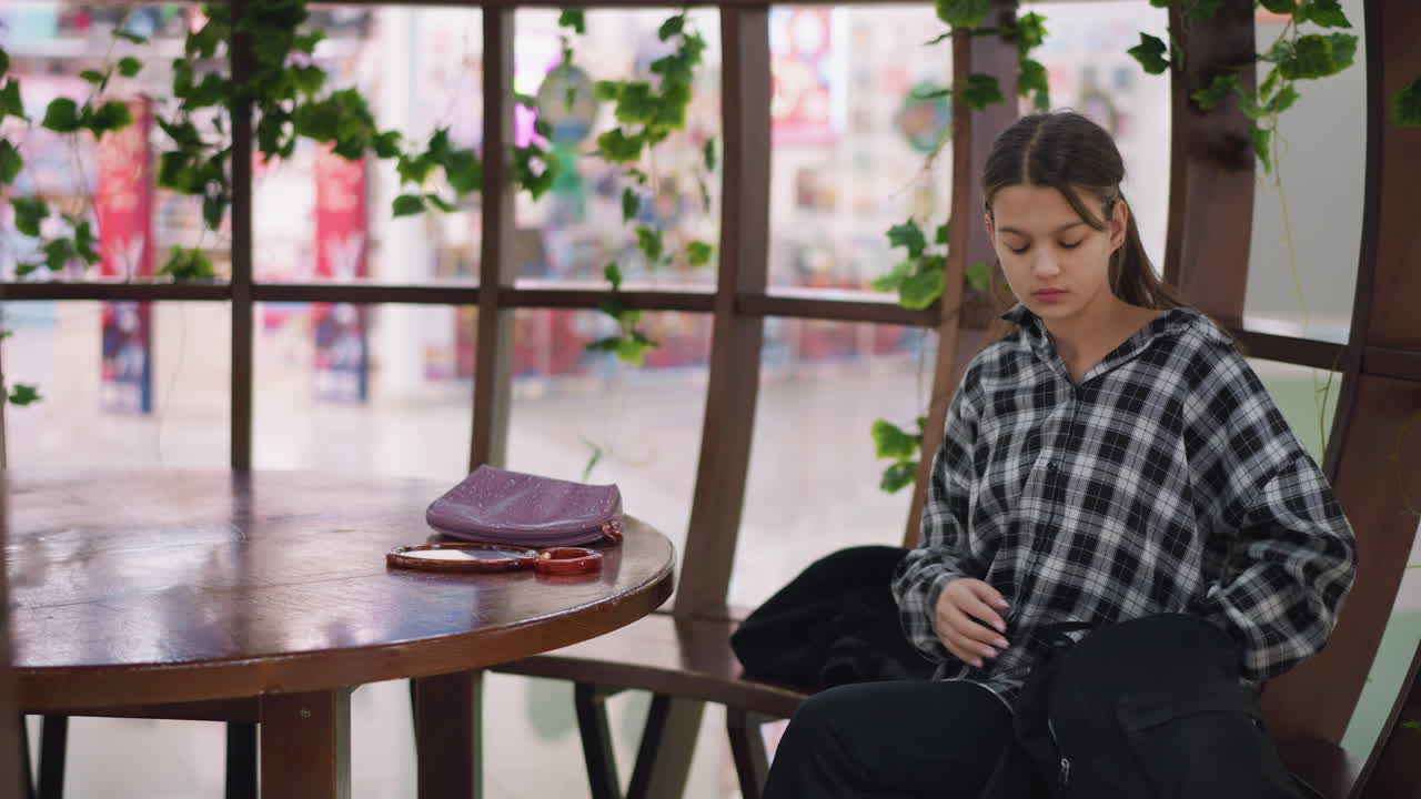 joven se sienta en una silla de madera, saca un espejo y un bolso de su bolsa, y examina su reflejo con una expresión enfocada y decidida en un espacio hermosamente decorado con vegetación