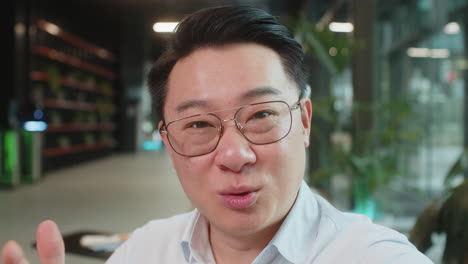 portrait of happy chinese businessman looking at camera and waves hand hello in office hotel lobby