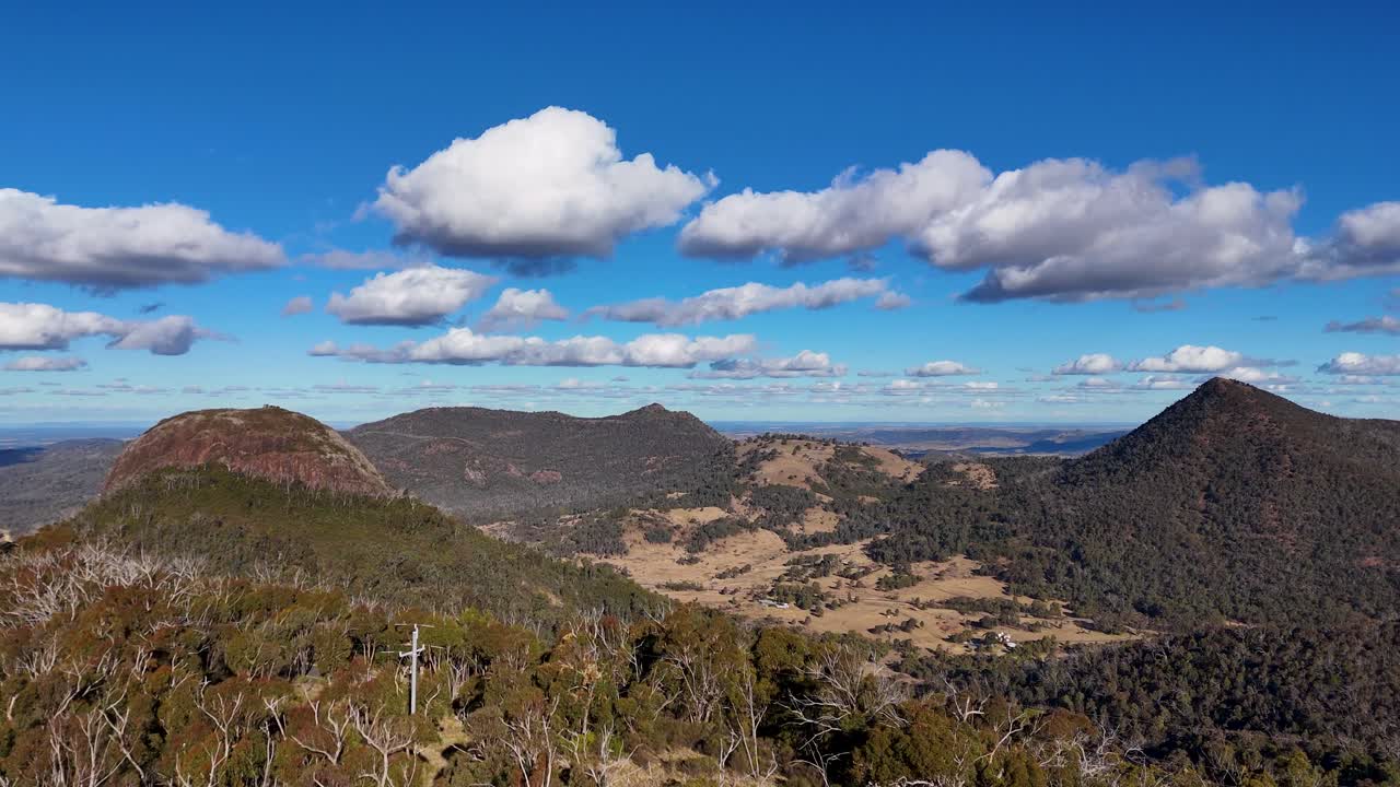 Aerial drone footage ascends above treetops to reveal expansive Warrumbungle National Park mountains under bright daylight, with smooth camera movement and clear blue skies