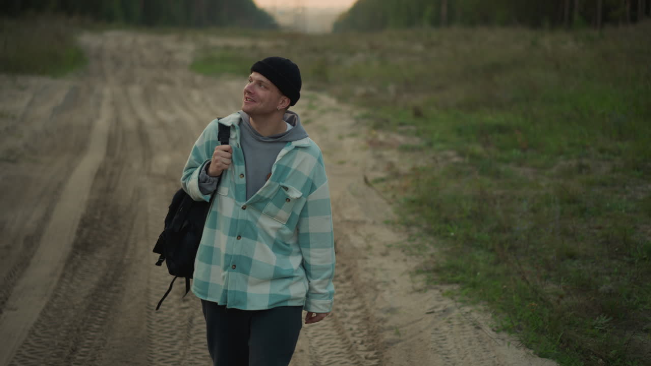 Escursionista caucasico su strada sterrata al crepuscolo, con giacca a quadri e berretto, che si ferma a studiare il cielo con uno zaino in spalla. Tranquillo sentiero nel bosco, luce soffusa cinematografica, viaggio in solitudine, atmosfera contemplativa