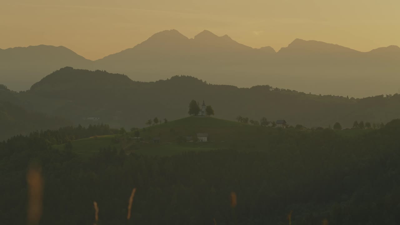 A couple with backpacks watches the sunrise over the mountains near St. Thomas Church, Slovenia