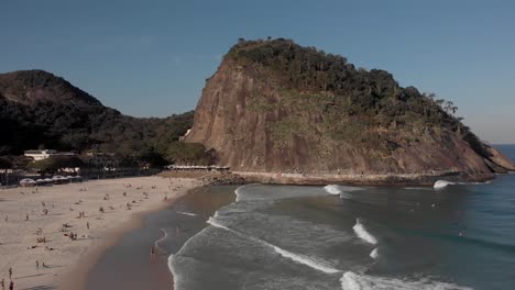 descenso aéreo que muestra a la gente en la playa de leme al final del bulevar de copacabana con la gran colina de rocas con el fuerte marcado por la bandera brasileña en la parte superior