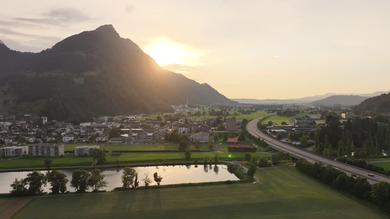 Luftfoto ved solnedgang over dalen i Glarus Nord, Sveits, med et boligområde og en liten innsjø som ligger under en fjellkjede.