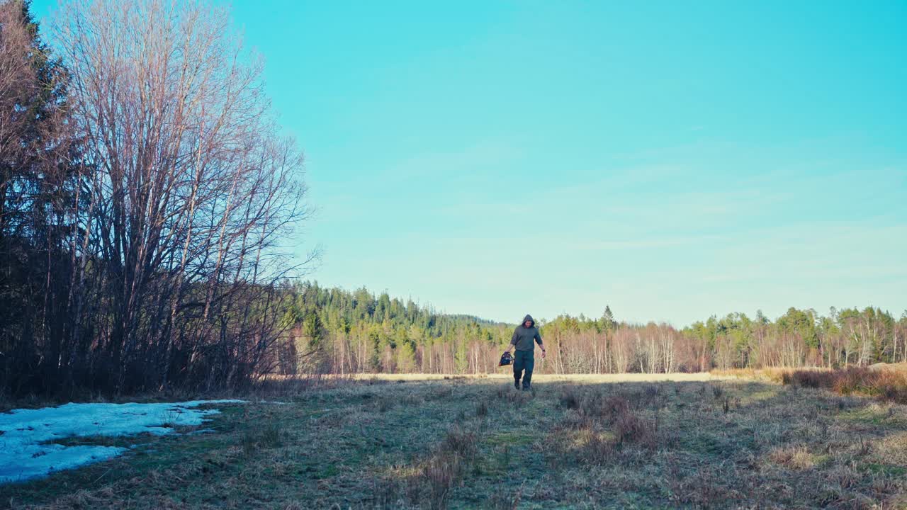 A Man Walks Through a Clearing at the Forest's Edge, Carrying a Chainsaw - Static Shot