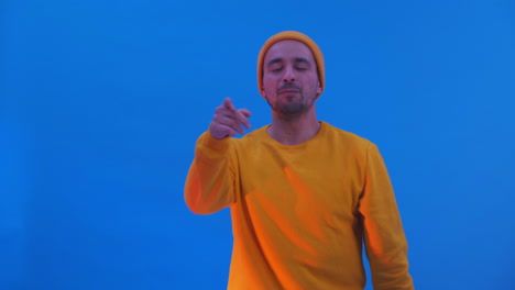 un hombre con capucha amarilla y sombrero está bailando sobre un fondo azul. baile divertido mientras mira a la cámara