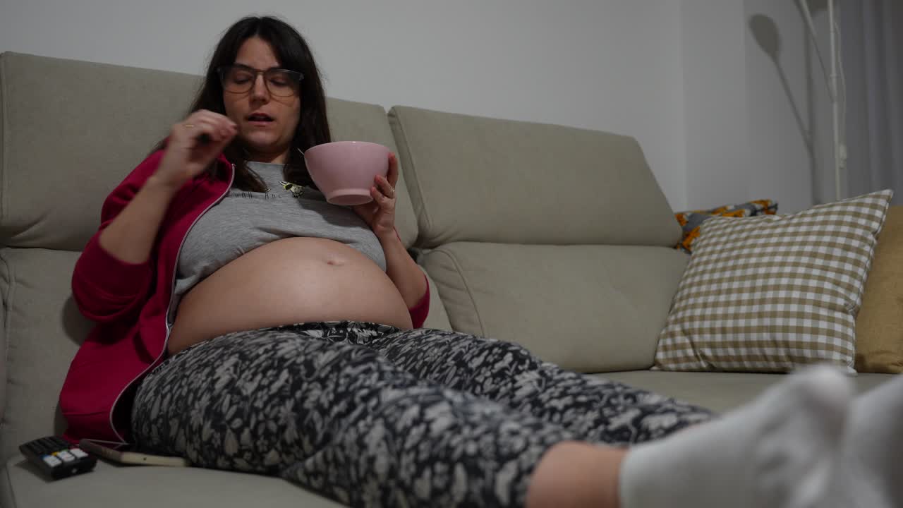 Pregnant woman on couch eating strawberries and using remote in cozy living room
