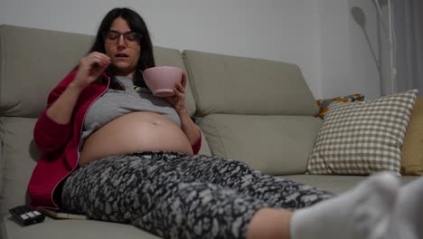 pregnant woman on couch eating strawberries and using remote in cozy living room