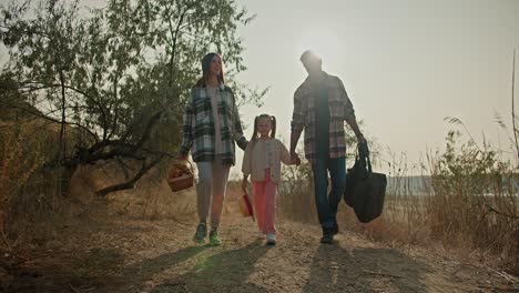 A-brunette-girl-in-a-Green-hat-in-a-Green-checkered-shirt,-along-with-a-basket,-walks-hand-in-hand-with-her-little-daughter-and-her-husband,-a-man-with-gray-hair-in-a-checkered-shirt,-on-a-picnic-in-the-summer