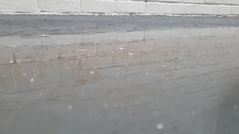 Rain-drops-falling-down-and-creating-ripples-on-a-concrete-slab-with-a-white-brick-wall-reflection-in-slow-motion,-sad-lonely-day-clip