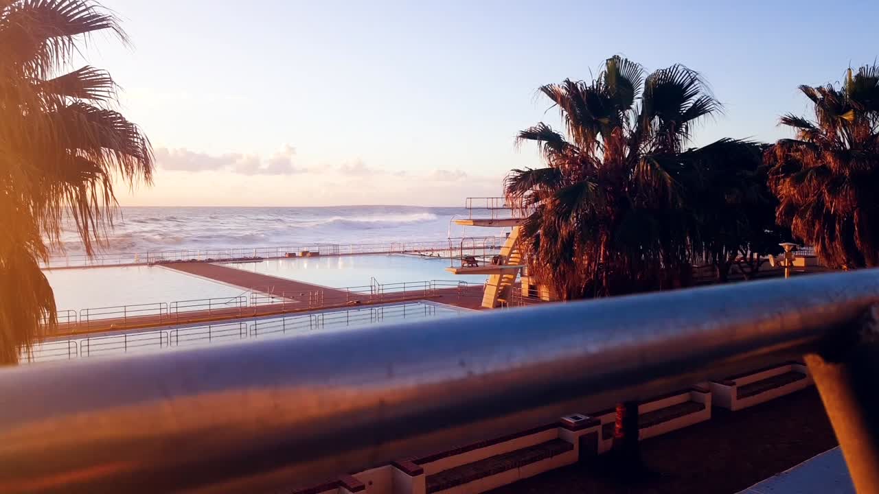 카메라가 난간 뒤에서 아래쪽으로 추적하는 일몰 동안 수영장과 바다