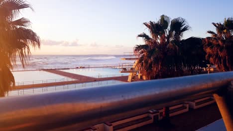 Badebecken-Und-Das-Meer-Bei-Sonnenuntergang,-Wobei-Die-Kamera-Hinter-Einem-Geländer-Nach-Unten-Fährt