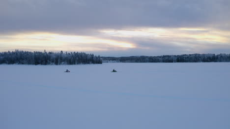 aerial view pair riding snowmobiles across flat snowy nordic winter wilderness at sunrise