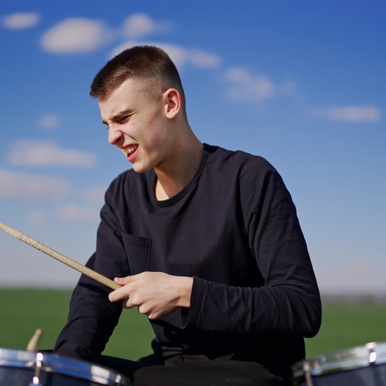 Jovem caucasiano tocando bateria expressivamente. Músico usa baquetas, golpeando a bateria ao ar livre com paixão. Vista em ângulo baixo