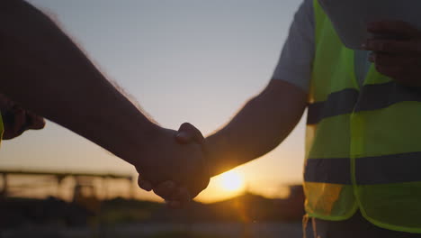 portrait of hands of two builders. builder shaking hand the builder on built house background. close up of a handshake of two men in green signal vests against the background of the sun