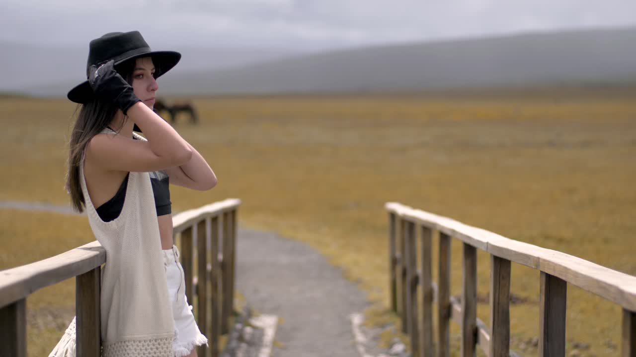 Woman wearing black gloves leans against wood railing on trail and puts on black hat