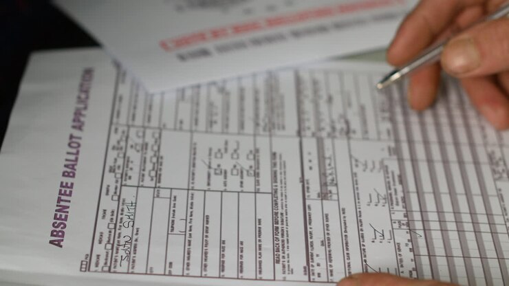 An absentee ballot application being filled out with a pen. An "Official Election Mail" envelope is visible in the background, emphasizing the mail-in voting process