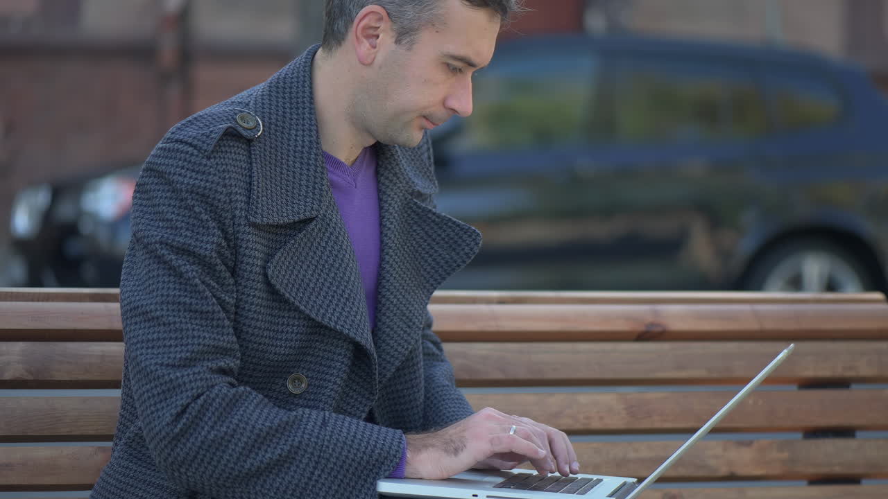 Man in a grey coat working on her laptop on a bench in the park
