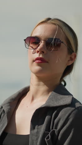 mujer blanca con camisa gris y gafas de sol sosteniendo bolsas de compras hacia atrás, mientras está de pie al aire libre con una expresión enfocada, el fondo está ligeramente borroso con personas parcialmente vistas