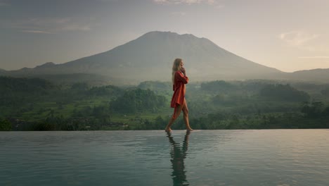 blond business woman in silk bathrobe walking on infinity pool edge while on phone call, mount agung in background