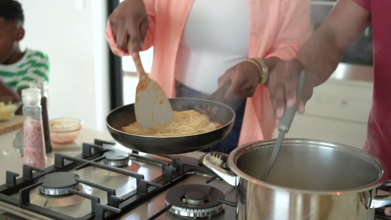 Família afro-americana preparando o jantar em ilha: mulher mexendo a panela e homem adicionando o macarrão após fervê-lo